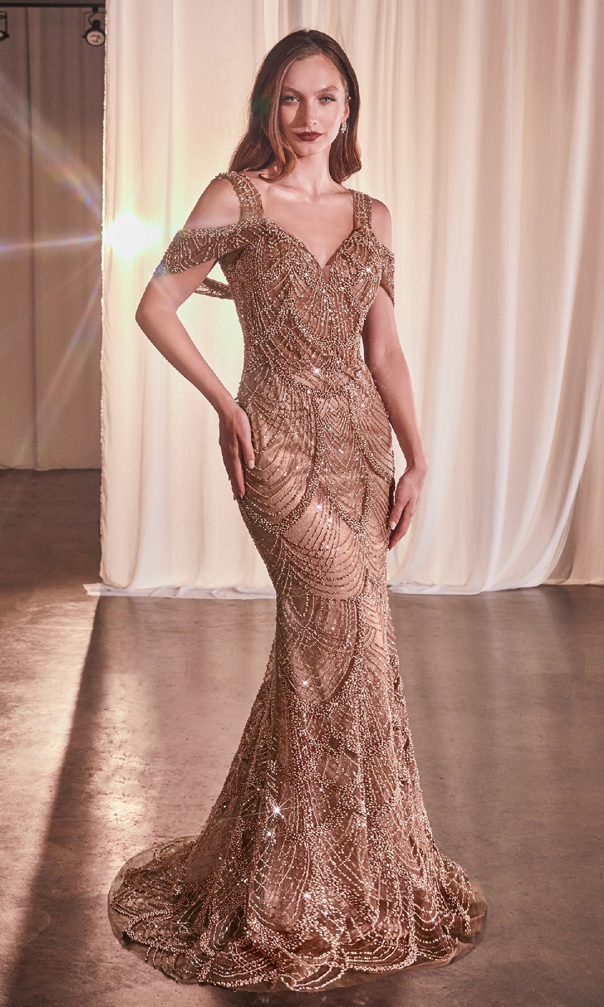 A woman in a long beaded sienna prom dress with a v-neckline and off-the-shoulder details, featuring a fully-lined long skirt with a sheer beaded overlay.